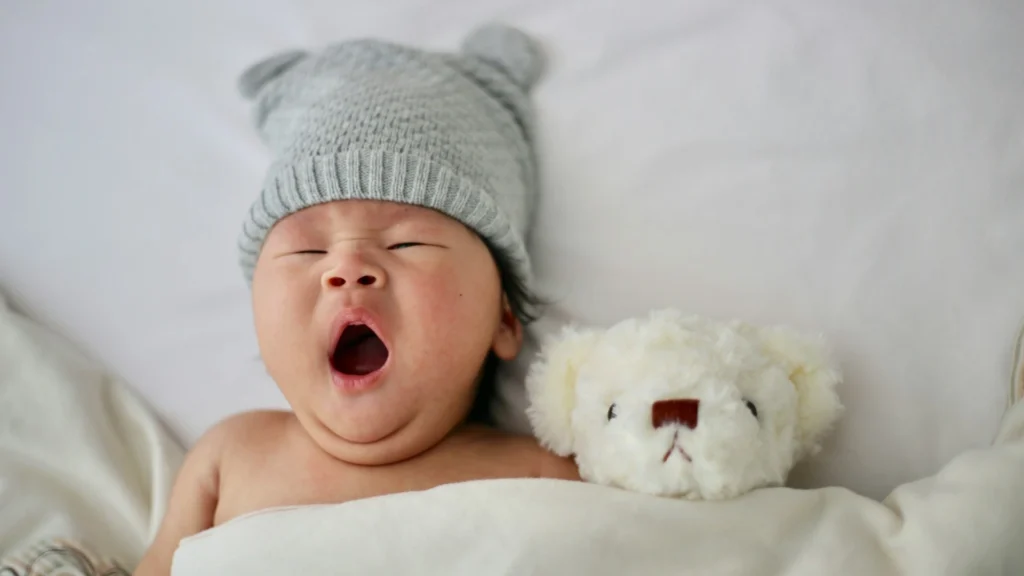 Yawning newborn baby wearing a gray knit hat and wrapped in a white blanket, lying next to a white teddy bear on a soft bed.