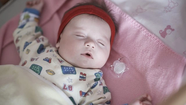 Baby peacefully sleeping on a pink blanket with a pacifier nearby, dressed in colorful pajamas and wearing a red headband, showcasing a calm and soothing sleep setting for infants.