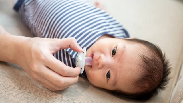 Newborn baby lying on a soft surface while a caregiver gently offers a pacifier, highlighting comfort and soothing techniques for infants.