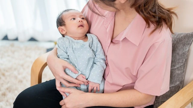 Mother holding a newborn baby dressed in a blue onesie with white patterns, sitting on a chair in a bright and cozy living room.