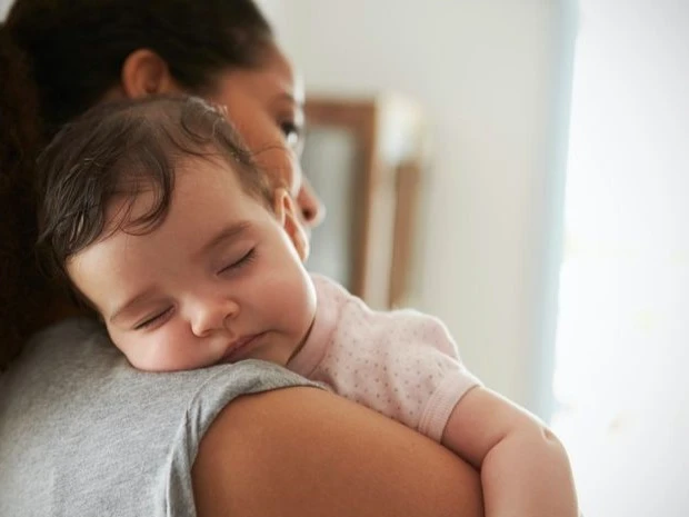 A mother holds her sleeping baby on her shoulder, providing warmth and comfort. The baby, dressed in a pink outfit, rests peacefully with a relaxed expression.