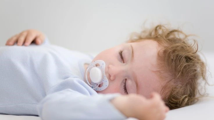 A peacefully sleeping baby with curly hair, wearing a light blue outfit, rests on a white surface while sucking on a pacifier for comfort.
