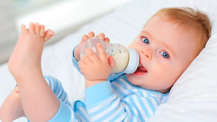 A baby with blue eyes and light brown hair, wearing a blue and white striped onesie, is lying on a white bed while drinking milk from a baby bottle. The baby is holding the bottle with both hands and has one leg playfully raised in the air. The baby looks content and relaxed.