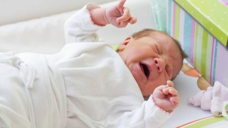 A newborn baby dressed in white is lying on a soft surface, crying with arms raised. The baby appears distressed, possibly due to discomfort or hunger.