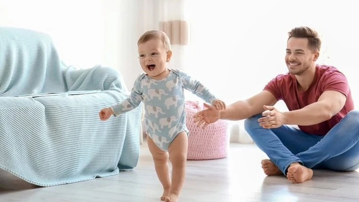 A joyful baby in a cute elephant-print onesie takes first steps toward a smiling dad, who sits on the floor with open arms, encouraging the milestone.