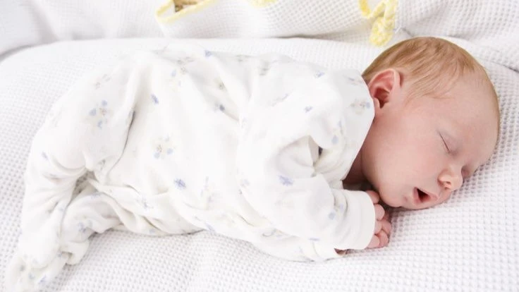 A newborn baby with light hair is peacefully sleeping on a soft white surface, dressed in a cozy white onesie with subtle patterns, resting in a curled position.