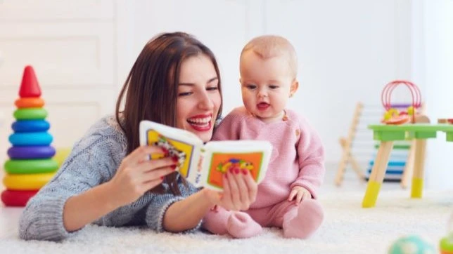 Mother reading a colorful picture book to her smiling baby on a soft carpeted floor, surrounded by toddler toys in a bright playroom.