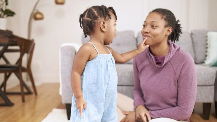 Toddler in a blue outfit touching her mother's lips while they engage in a playful and interactive moment on the floor of a cozy living room.