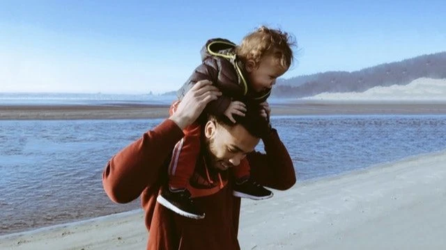 A father carrying his young child on his shoulders while walking along a beach with a scenic backdrop.