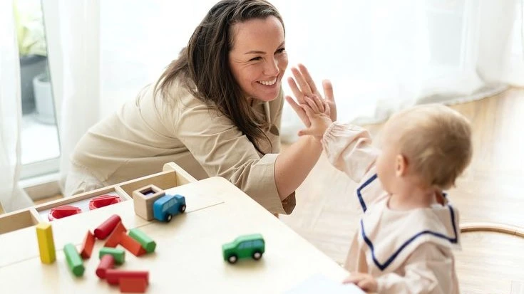 A smiling mother high-fiving her baby at a play table with colorful wooden toys, reinforcing positive parenting and child development.