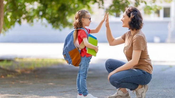 A mother crouching down and giving her young daughter a high-five before school, promoting confidence and positive reinforcement in parenting