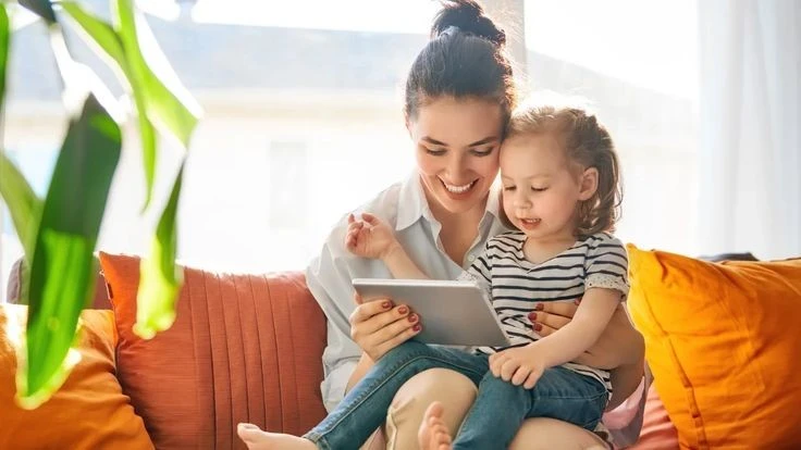 A smiling mother and her young daughter sitting on an orange couch, happily looking at a tablet together in a bright and cozy living room.