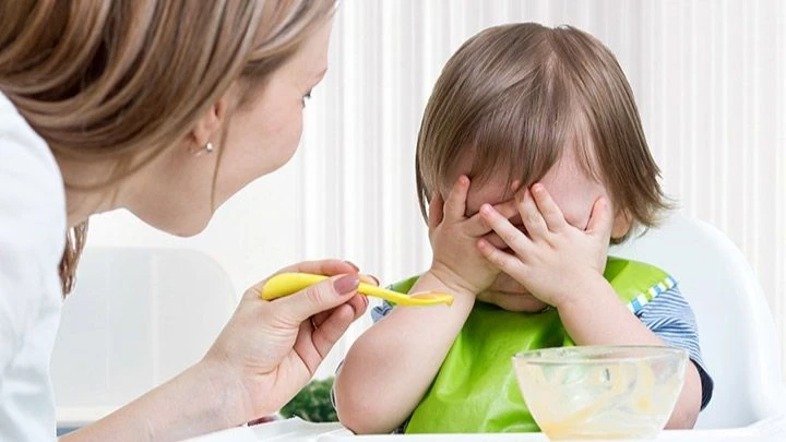  Frustrated toddler covering face while being fed, highlighting challenges of picky eating and toddler meal prep struggles.