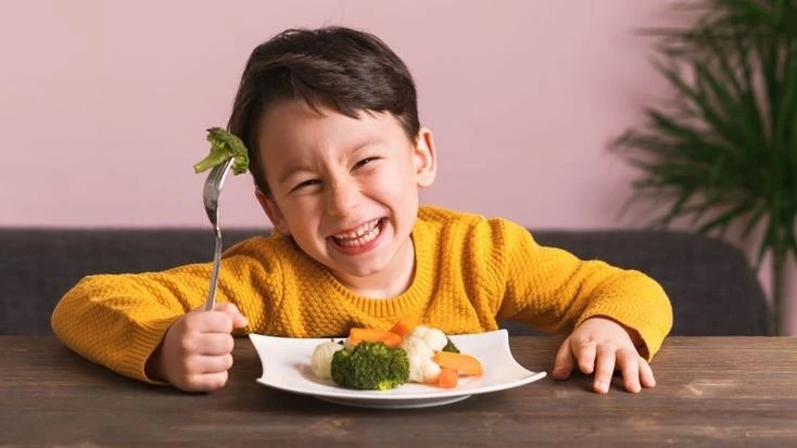 Smiling toddler holding a fork with broccoli while enjoying colorful vegetables, showcasing joyful toddler meal prep ideas.